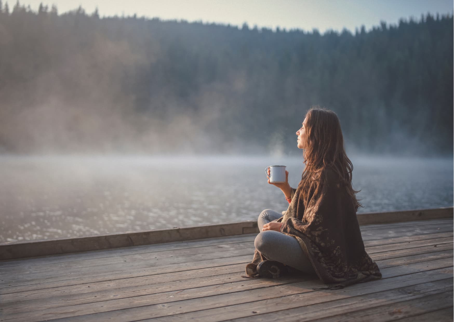 Kobieta siedzi na pomoście nad brzegiem jeziora z kubkiem gorącego napoju.