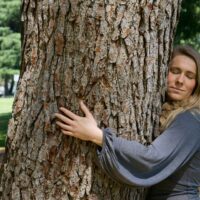 Kobieta przytula się do drzewa a na jej twarzy widoczna jest radość i spokój.