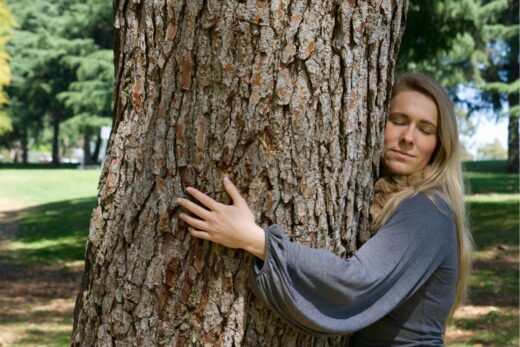 Kobieta przytula się do drzewa a na jej twarzy widoczna jest radość i spokój.